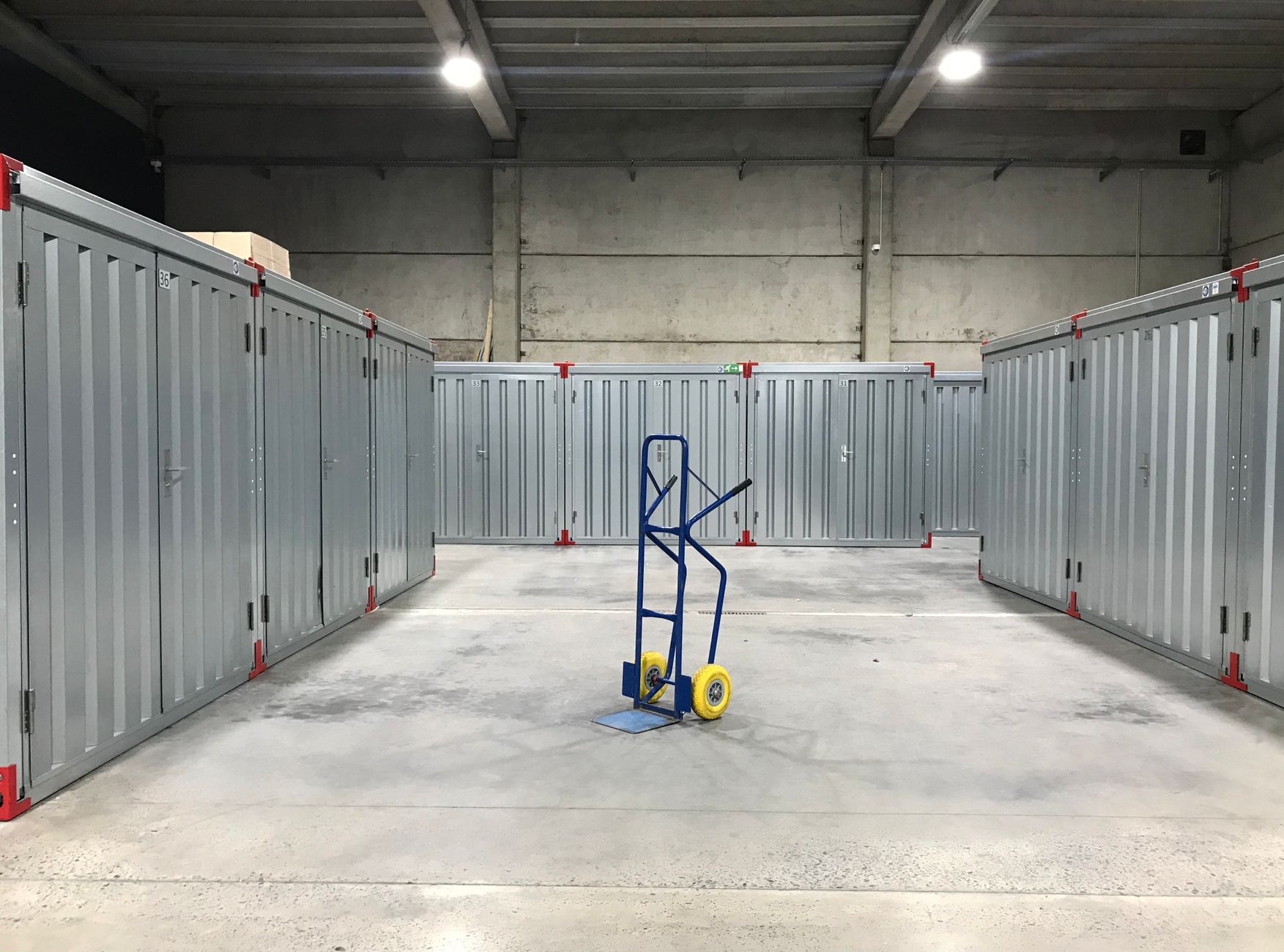allée de box de stockage métalliques numérotés dans un entrepôt, avec un diable au centre